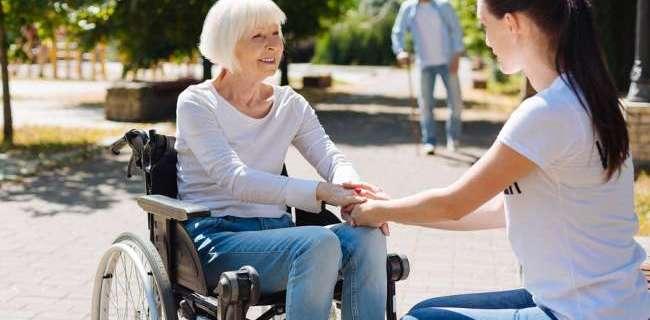 Quick recovery. Lively graceful aged lady sitting in a wheelchair and recovering from leg injury while young woman providing her with psychological support